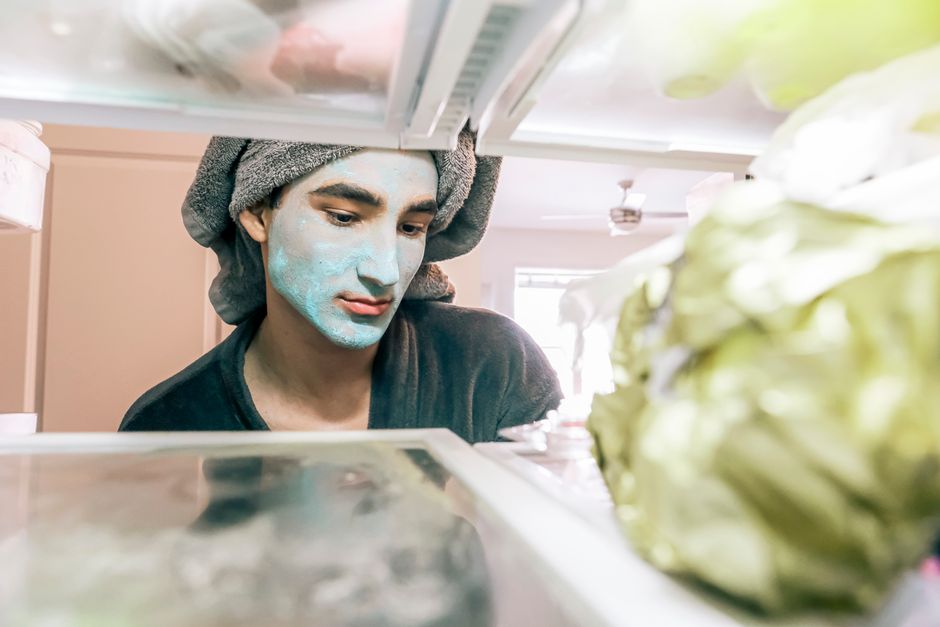 A man with a blue face mask looks into the refrigerator, towel wrapped on head, in a brigh