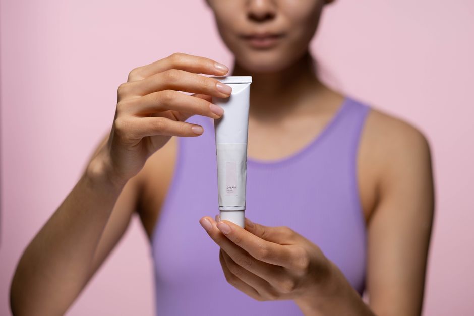 A woman in a purple tank top holds a skincare cream against a pink background in a studio 