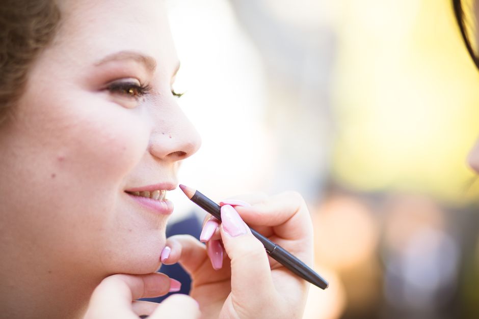 A woman smiling as a makeup artist applies lip liner outdoors in soft daylight.