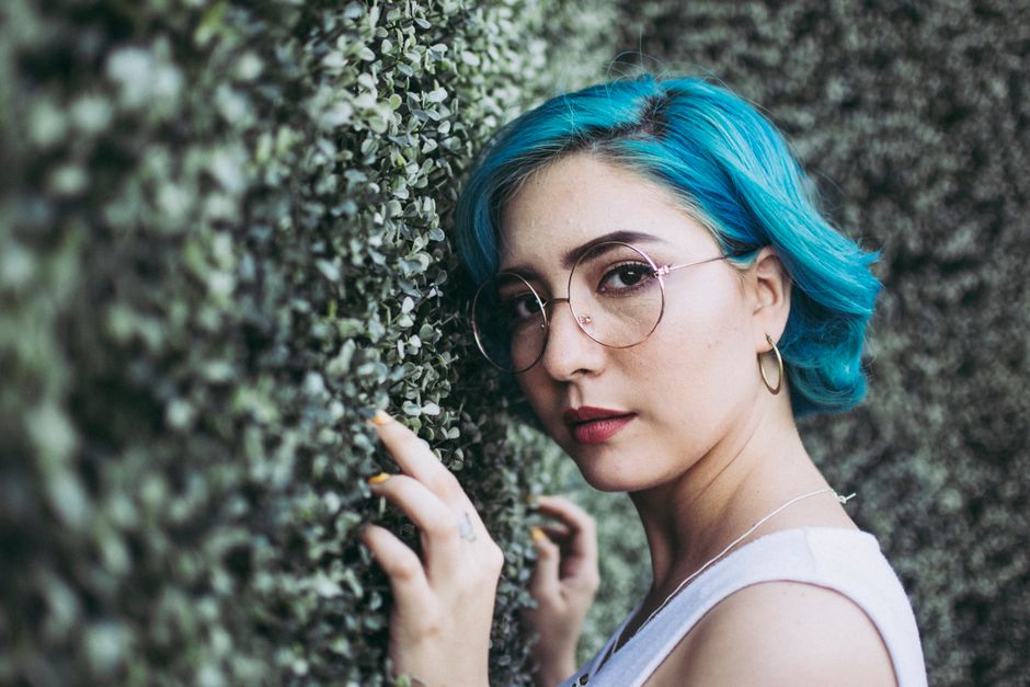 Fashionable woman with blue hair and glasses posing against a leafy backdrop outdoors.