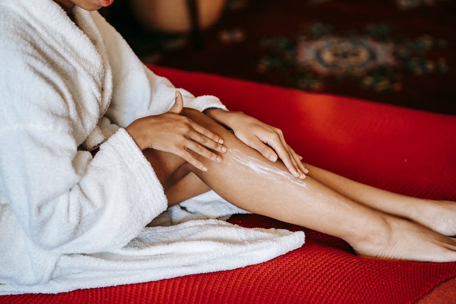 Faceless female in bathrobe applying lotion on leg with delicate skin while sitting on red sofa