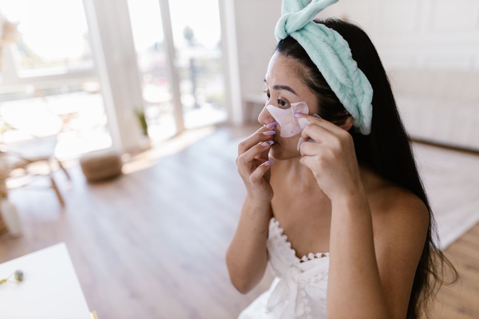 Young woman with long hair applying eye patches in a cozy indoor setting.