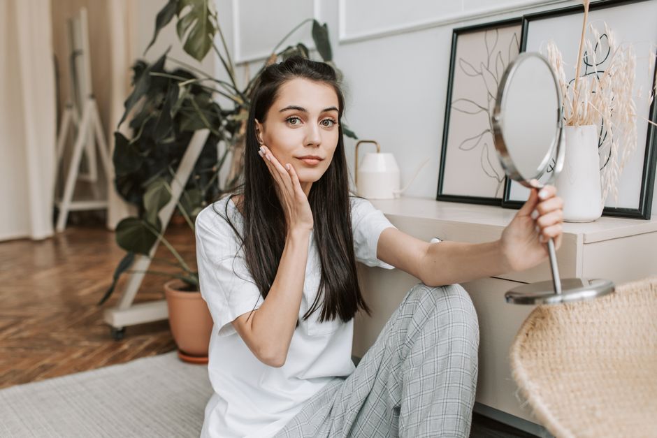 Woman sitting in living room using mirror for facial care routine. Indoor setting.