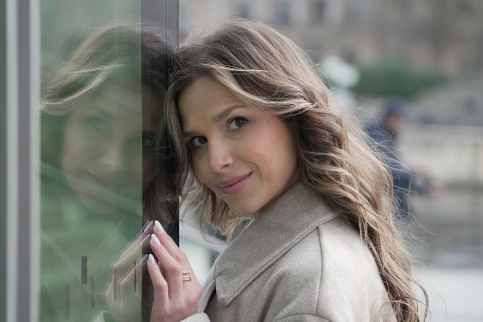 A fashionable woman with brown hair smiling near a window with her reflection in Berlin, Germany.