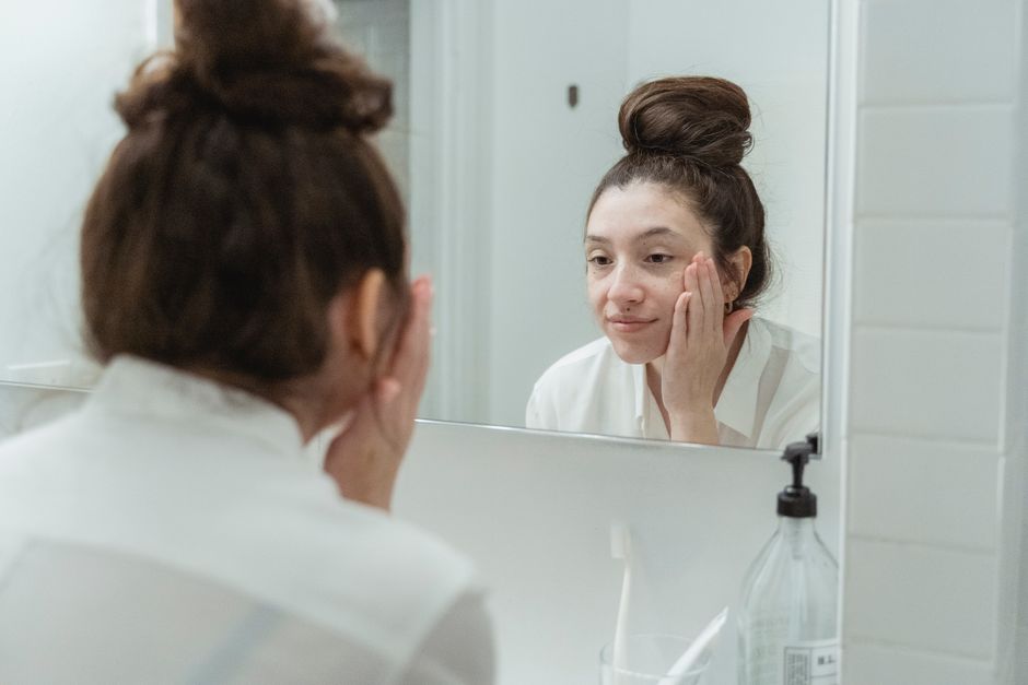 Woman looking at her reflection in the mirror while applying skincare products.