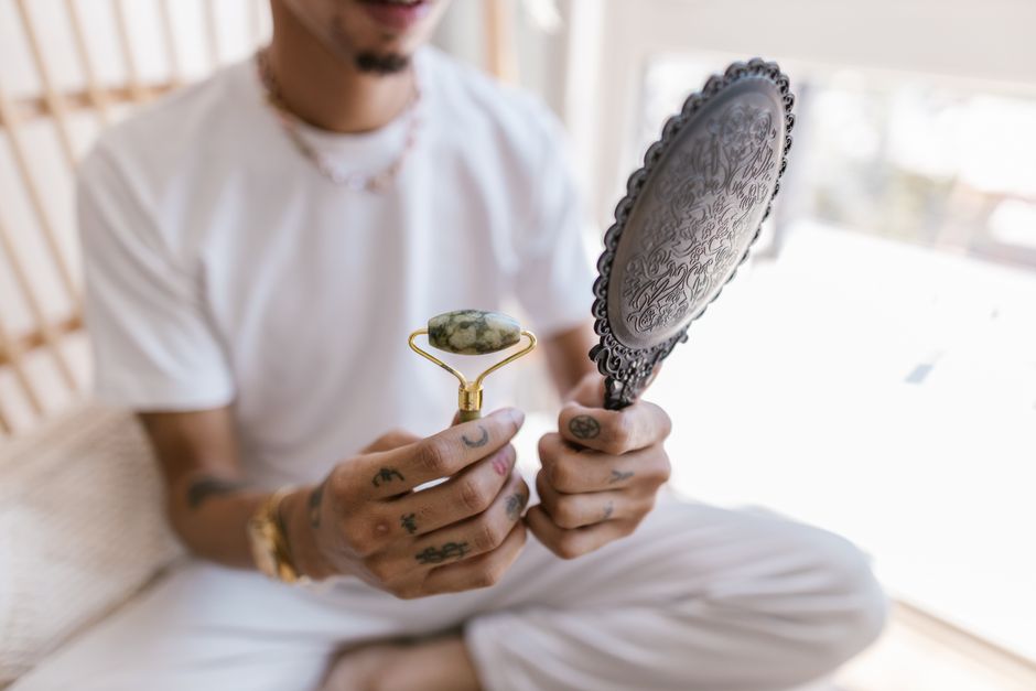 A tattooed man holds a jade roller and ornate mirror in a bright setting.