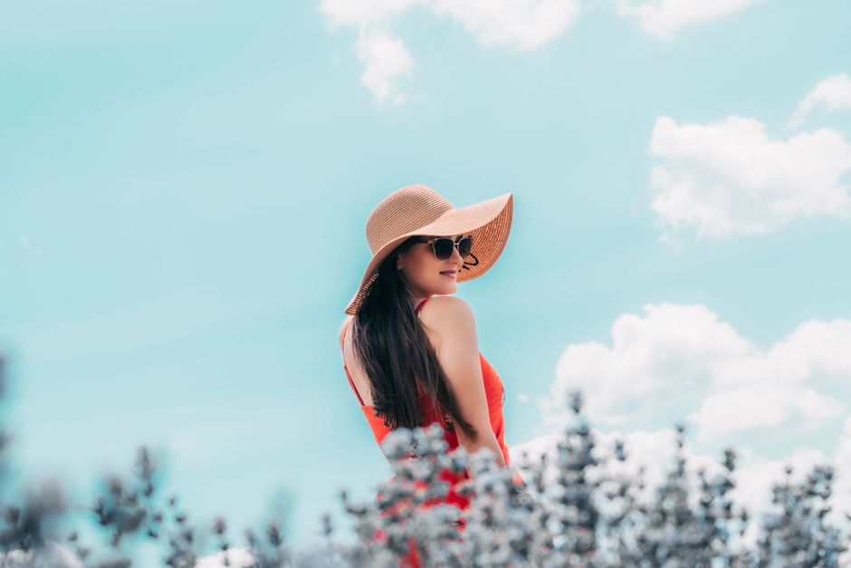 Back view young content brunette wearing trendy hat with wide brims and sunglasses standing near blurred flowering plants against cloudless blue sky