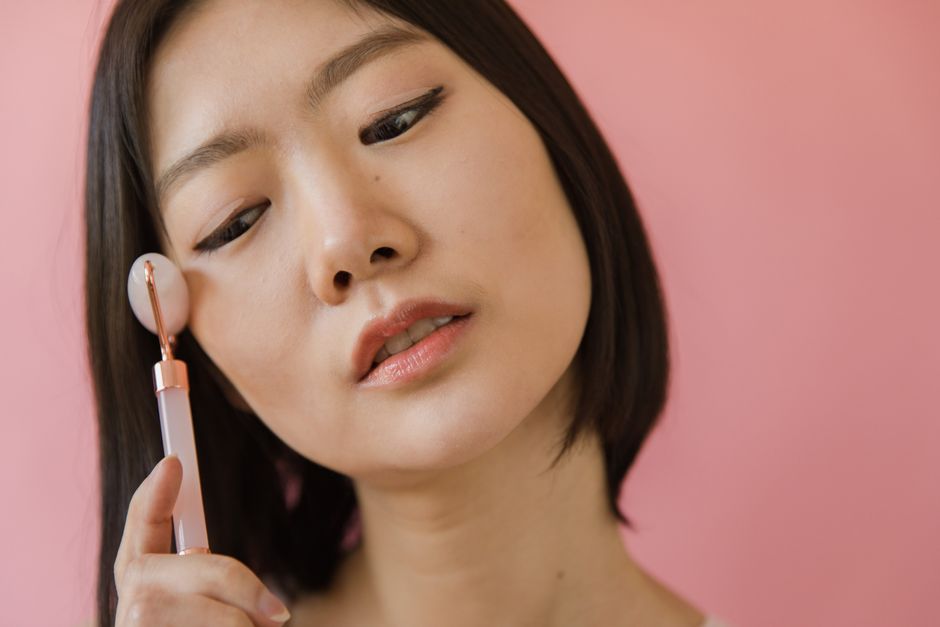 Close-up of an Asian woman using a jade roller for facial massage against a pink backgroun