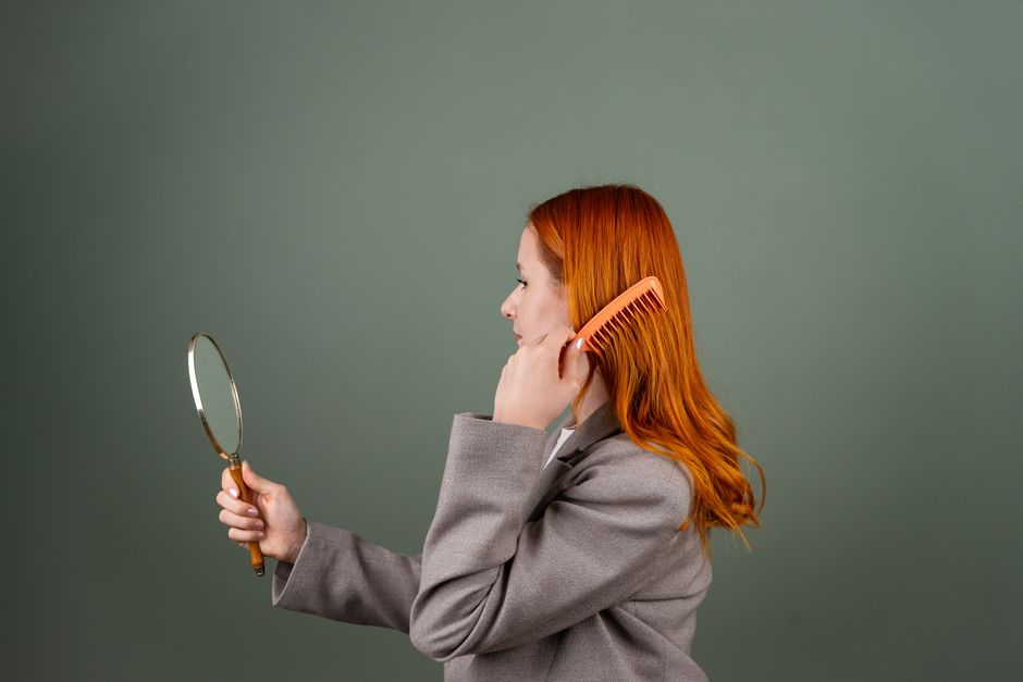 Side profile of a woman combing her hair and looking in a handheld mirror.
