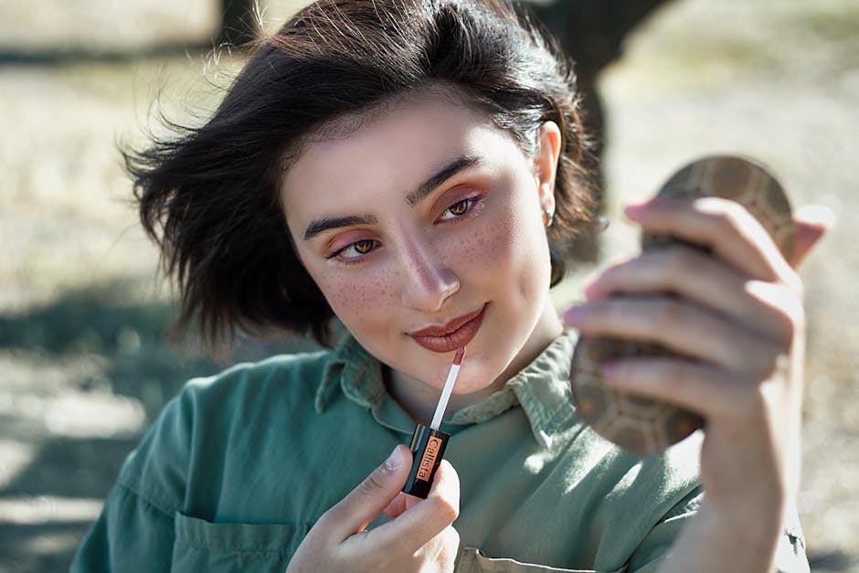 A young woman with freckles applying lipstick using a pocket mirror outdoors.