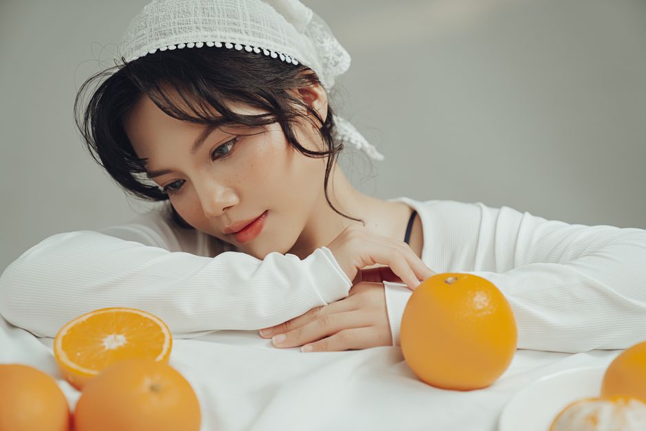 Thoughtful woman wearing white with oranges on table, serene indoor scene.