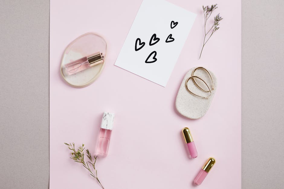 Elegant flat lay of perfume, lip gloss, and card with hearts on a pink background.