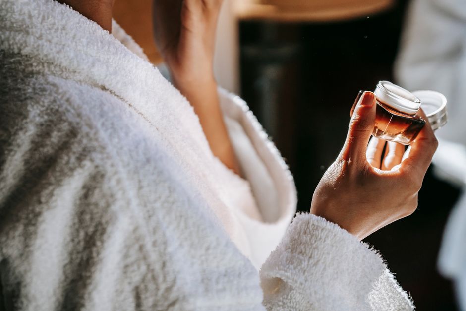 Side view of crop anonymous ethnic female with jar of moisturizing cream against mirror du