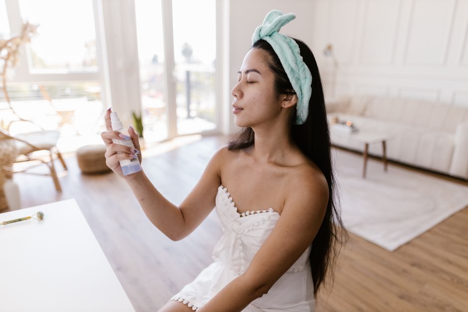 Woman sprays face mist for skincare in a cozy living room with natural light.