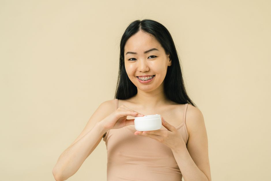 Asian woman with braces smiling while holding a cosmetic jar in a studio setting, representing skincare and self-care.
