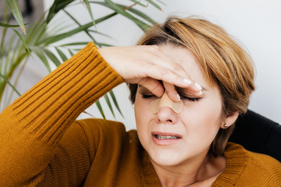 Woman holding her nose with a band-aid, showing discomfort and pain indoors.